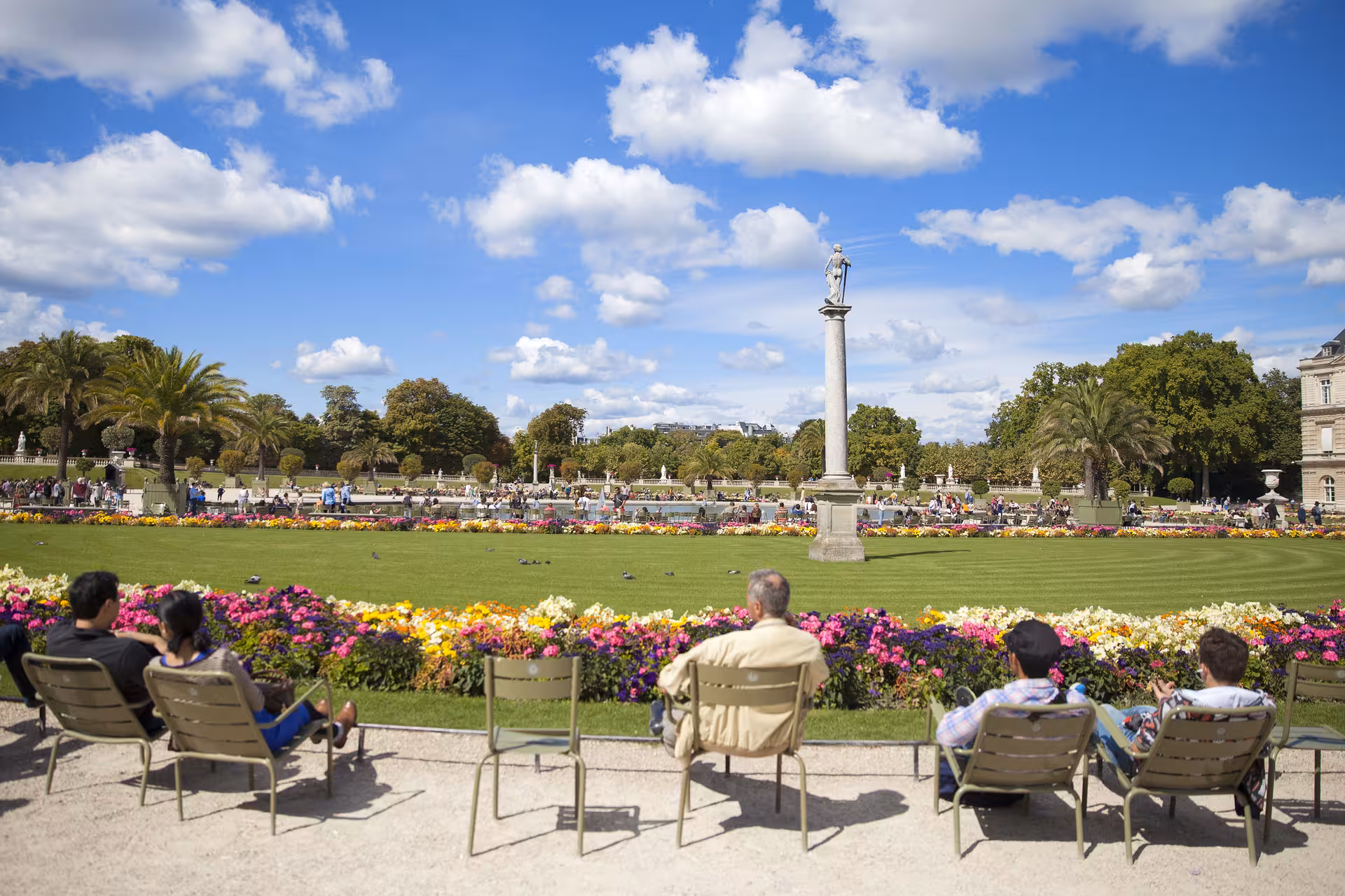 Visitors relaxing in chairs at Jardin du Luxembourg, Paris, with colorful flowers and a stone column statue