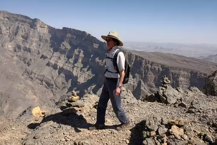 Hiker with sunhat and backpack stands on rocky ledge overlooking rugged Jebel Shams canyon during guided balcony walk.
