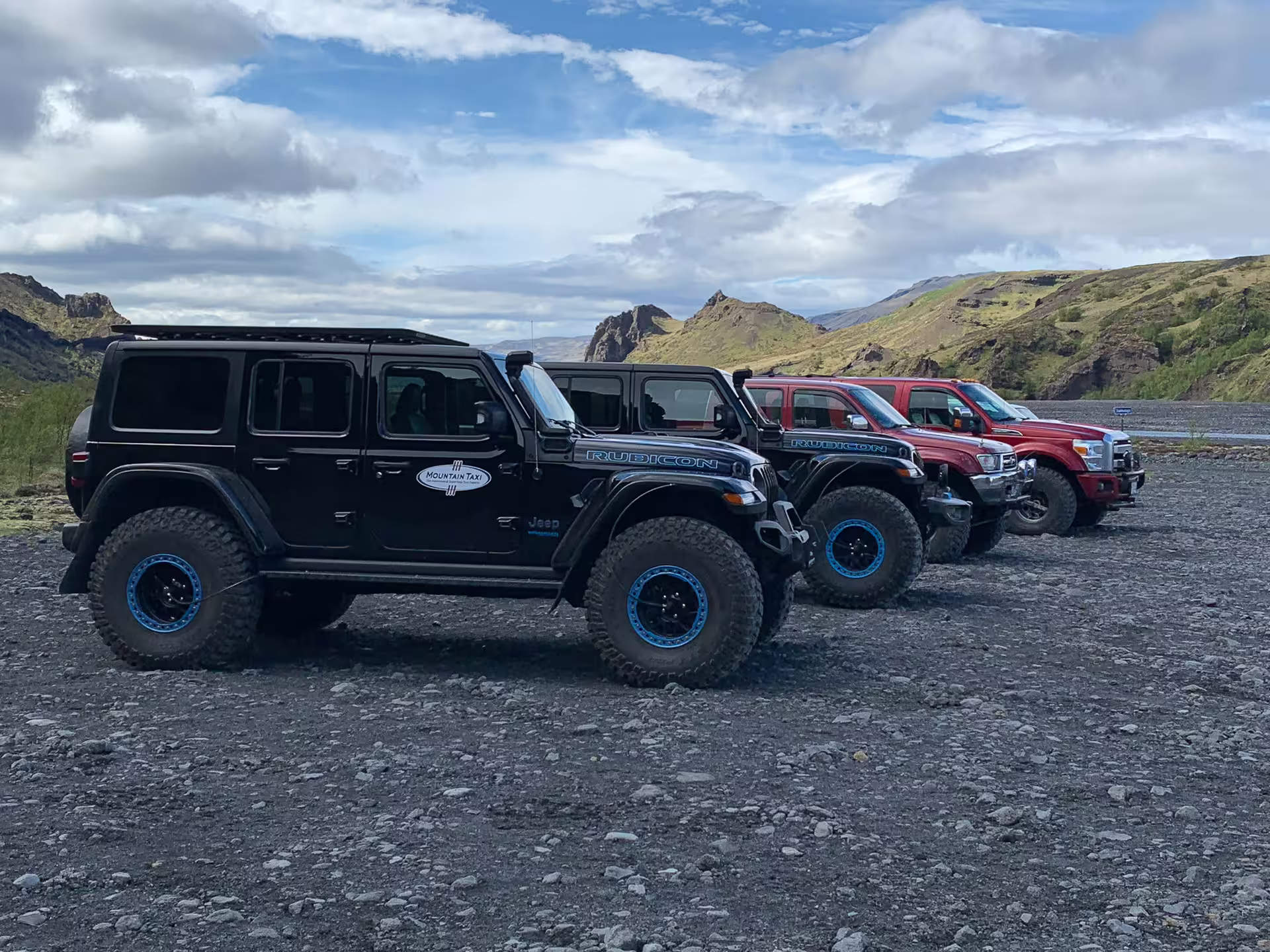 Fleet of black Jeep Wrangler Rubicon 4x4 vehicles parked on volcanic gravel with Iceland mountains behind