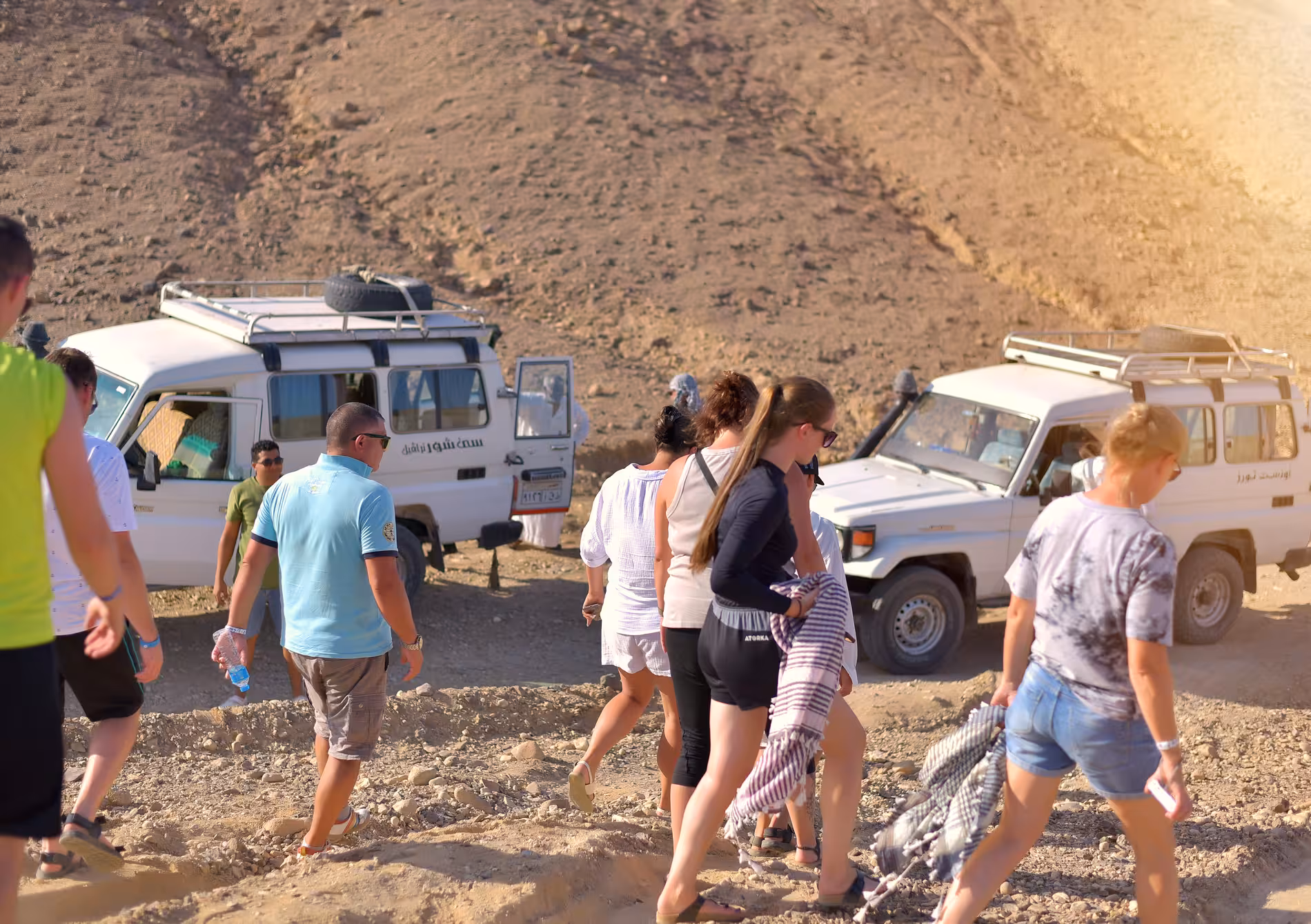 Tourists walking near white 4x4 jeeps in rocky desert landscape during safari in Marsa Alam