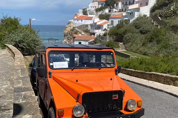 Experience the scenic coastal drive to Cascais in a classic orange jeep with ocean views.
