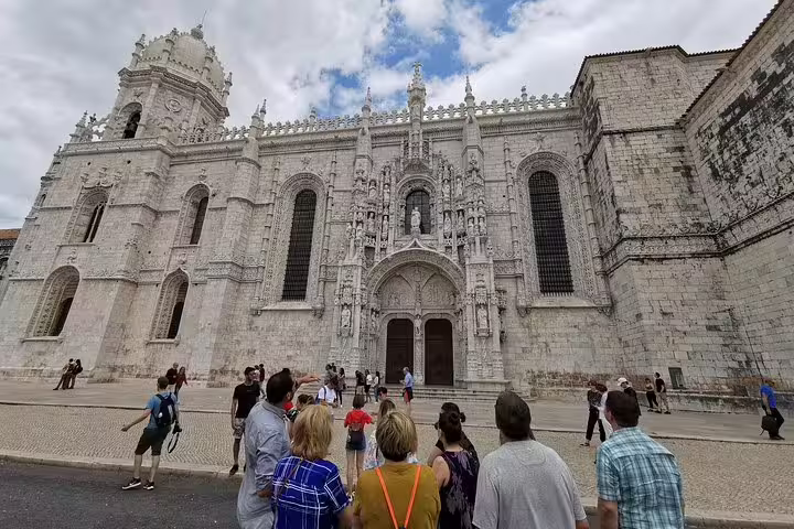 Tourists admire the grand entrance of the Jerónimos Monastery, a highlight on the Porto to Lisbon transfer with stops in Nazaré, Alcobaça, and Óbidos.