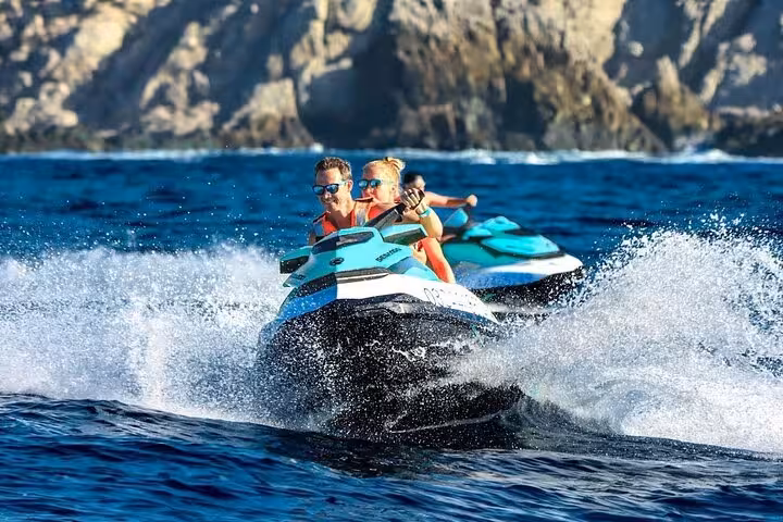 Two people riding a turquoise jet ski at speed near rocky coastline in Golfe-Juan