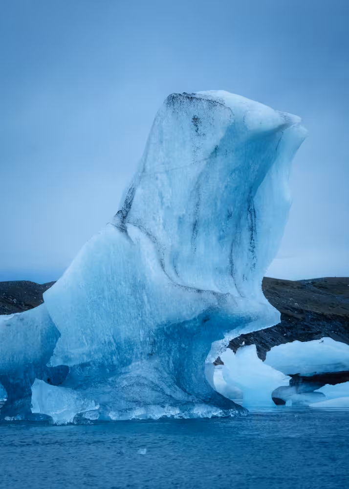Towering blue iceberg floating on Jokulsarlon glacial lagoon with dark rocky shores in background
