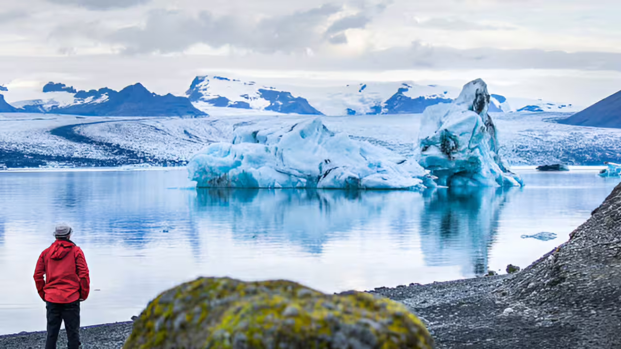 Traveler in red jacket gazing at blue icebergs floating on Jokulsarlon Glacier Lagoon, Iceland