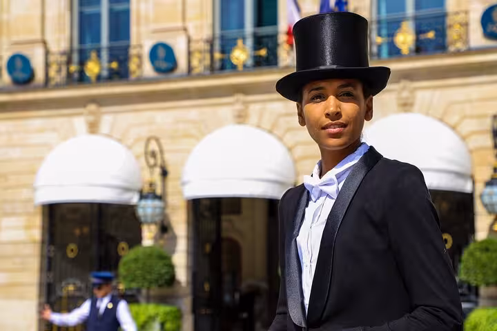 Elegantly dressed performer in black tuxedo and top hat stands before a grand Parisian hotel facade