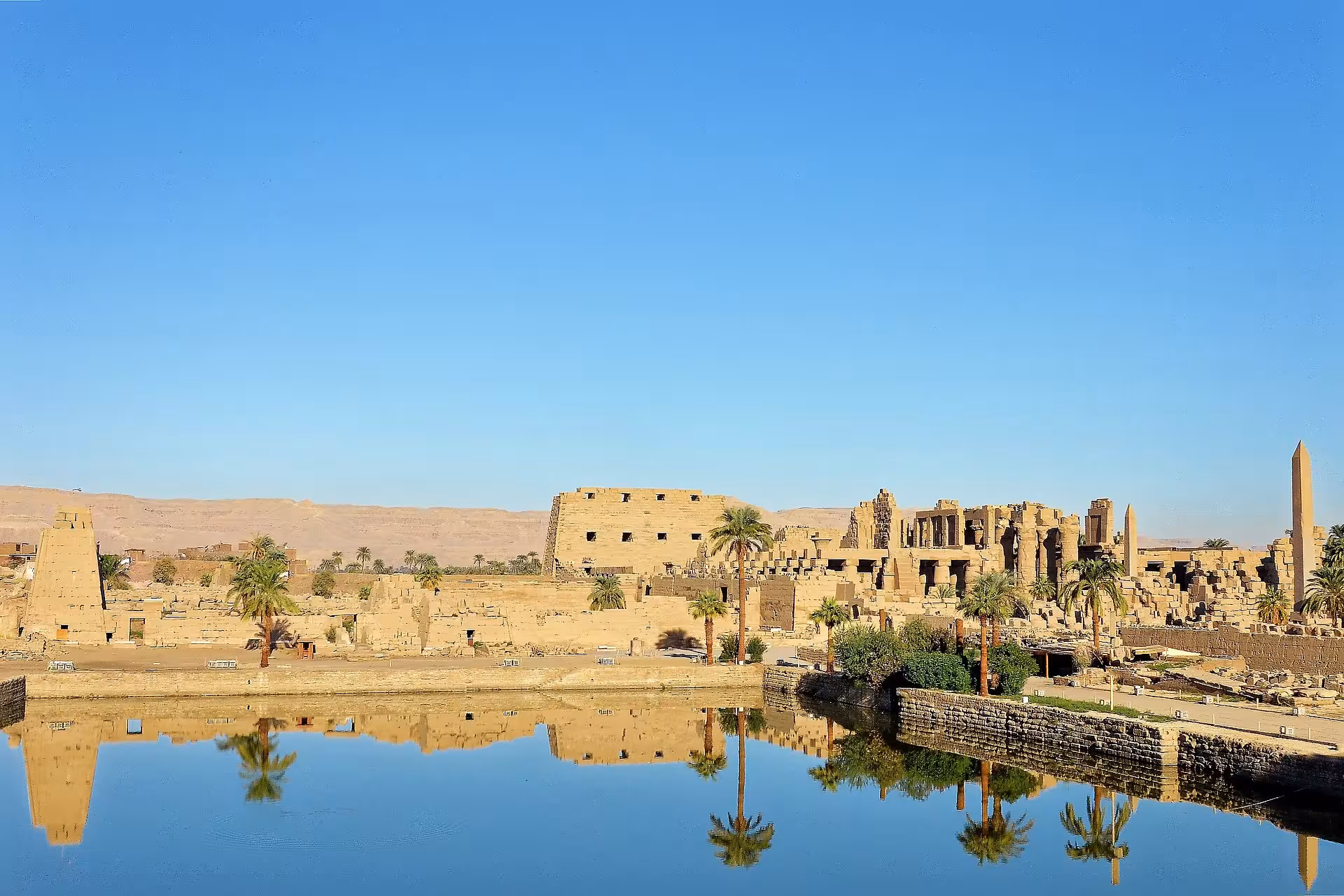 Karnak Temple complex reflected in sacred lake, Luxor, Egypt, with obelisks and palm trees under clear blue sky