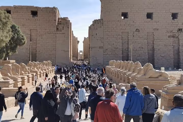 Tourists exploring the grand entrance of Karnak Temple, lined with sphinx statues, under a clear blue sky.