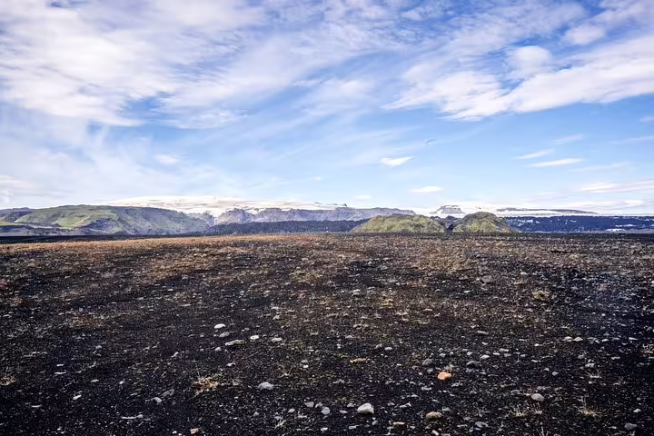 Black volcanic lava field with snow-capped mountains and green hills near Katla, Iceland