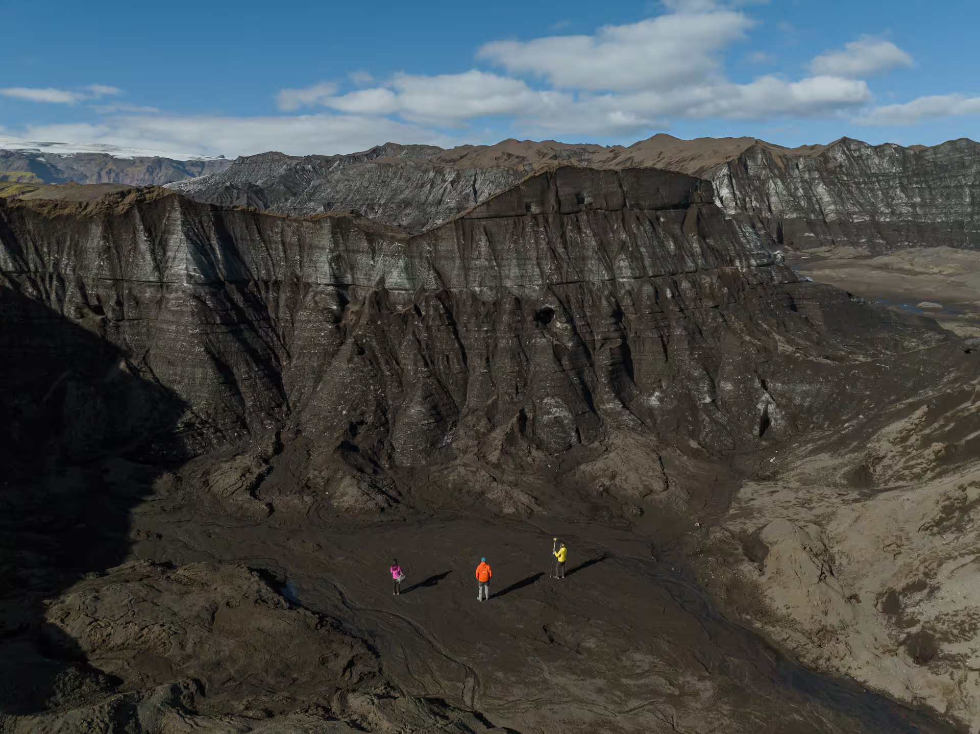 Three hikers standing before massive ash-covered dead ice cliffs on a full-day Katla Glacier guided tour in Iceland