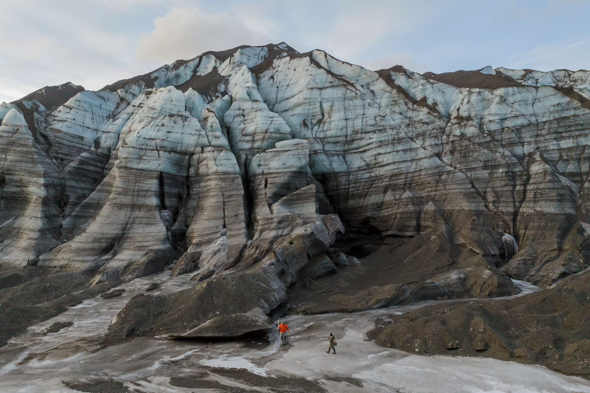 Two explorers stand before the massive blue-streaked ice walls of Katla glacier on a guided research tour in Iceland.