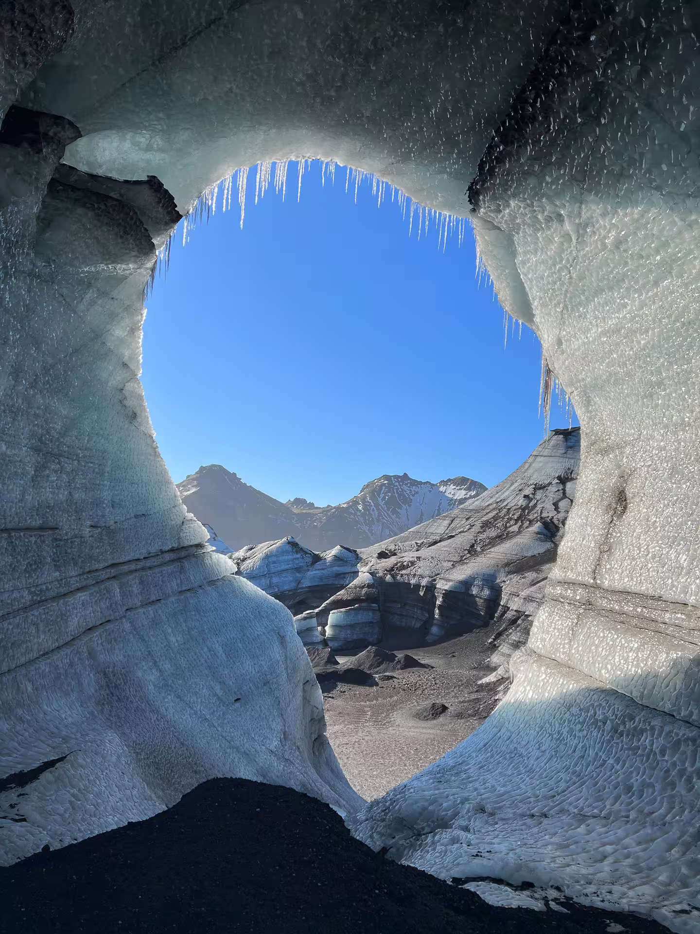View through a glacial ice cave arch with icicles framing snow-capped Icelandic mountains under blue sky