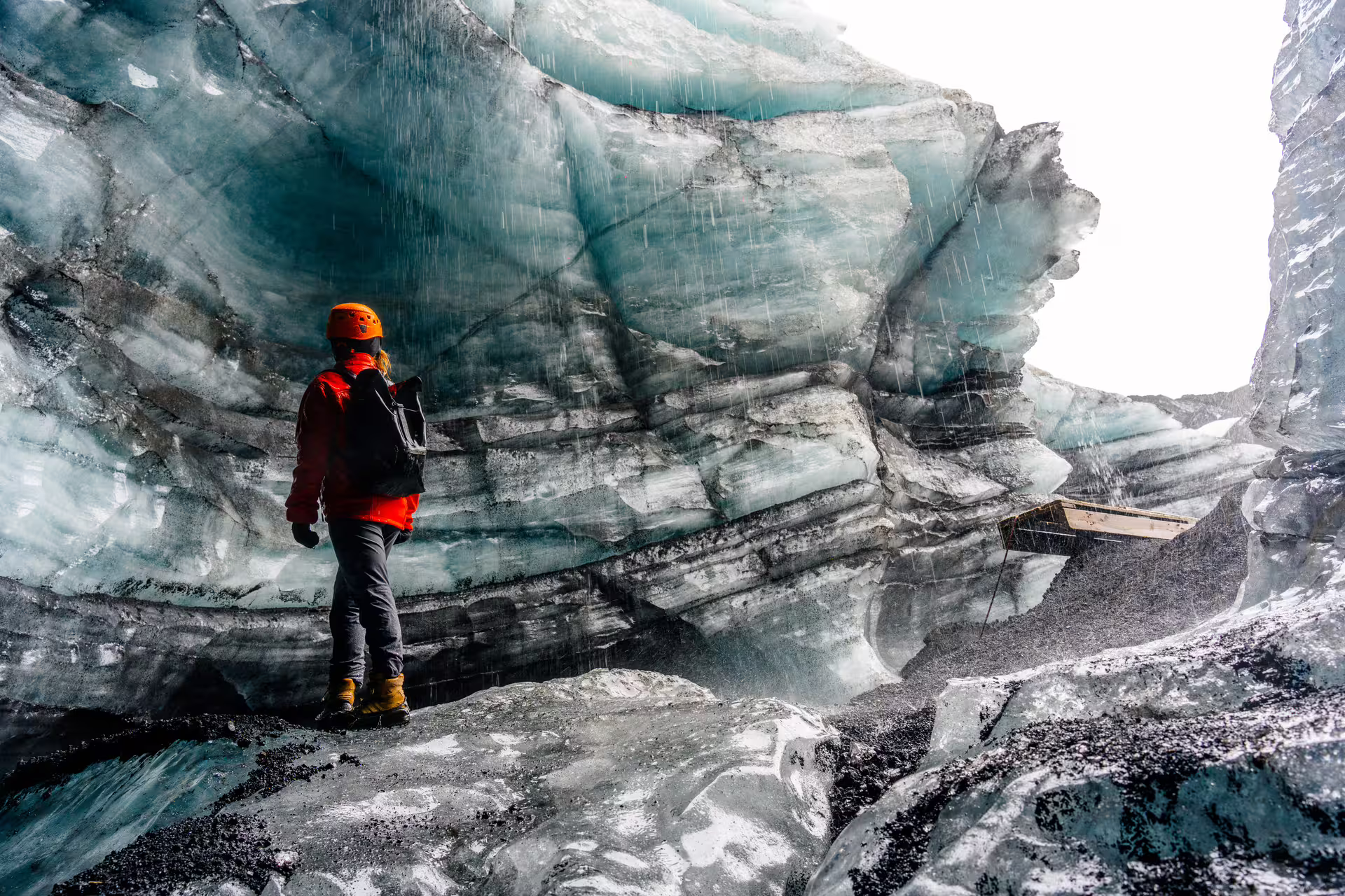 Hiker in red jacket and orange helmet exploring Katla ice cave with layered glacier walls