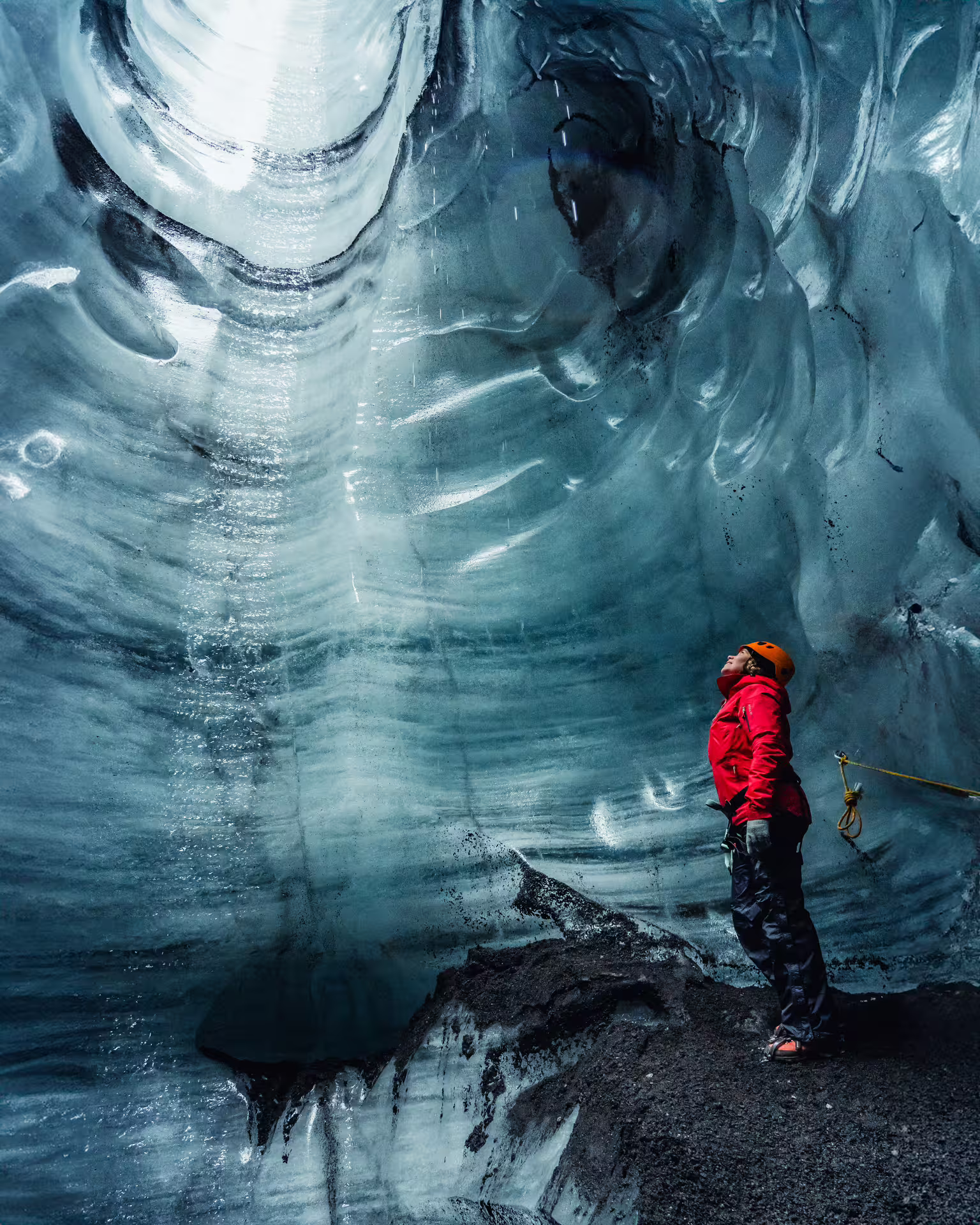 Explorer in red jacket and helmet gazing up at swirling blue ice walls inside Katla ice cave