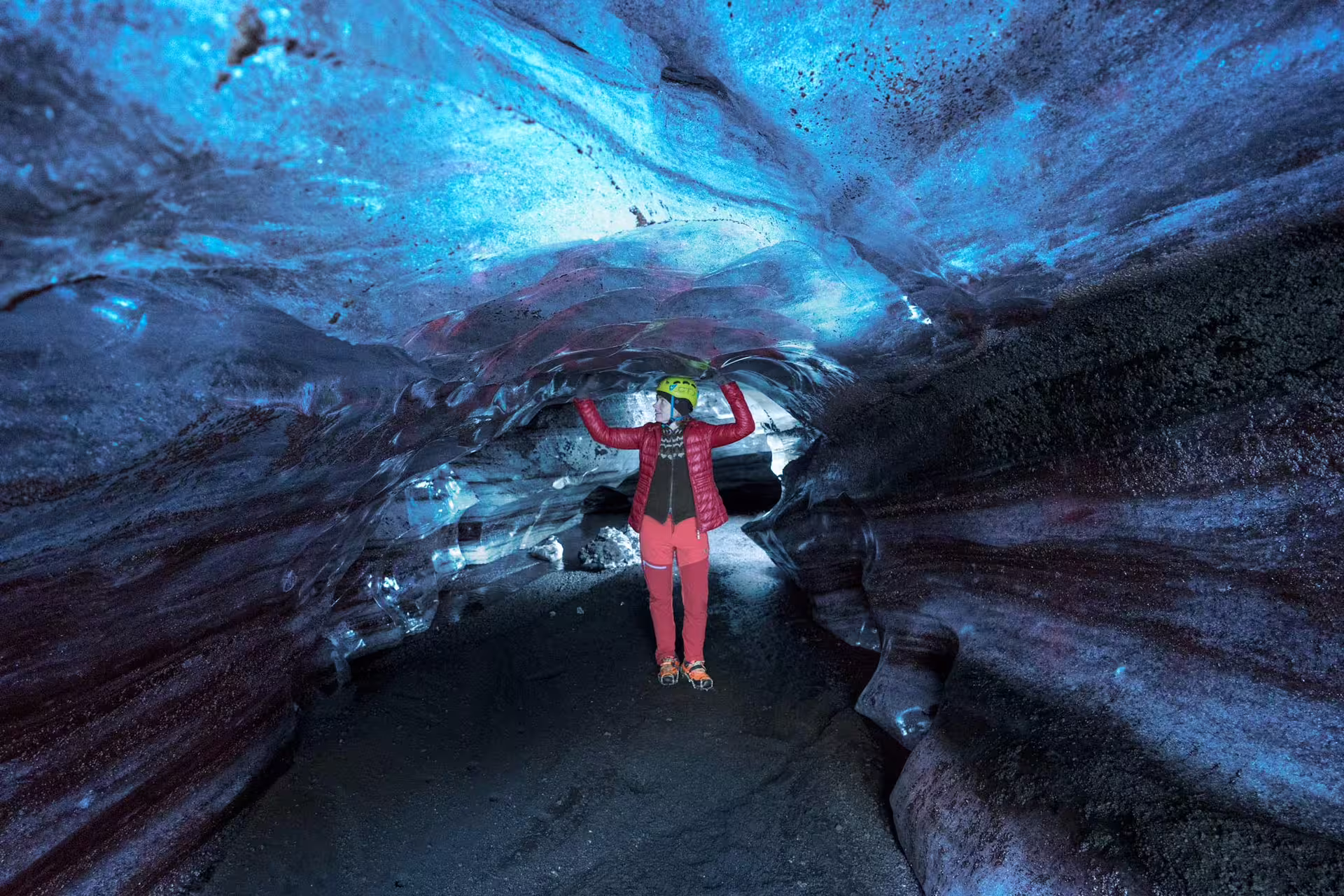 Explorer in red jacket and helmet touching ice ceiling inside Katla ice cave with glowing blue glacier walls