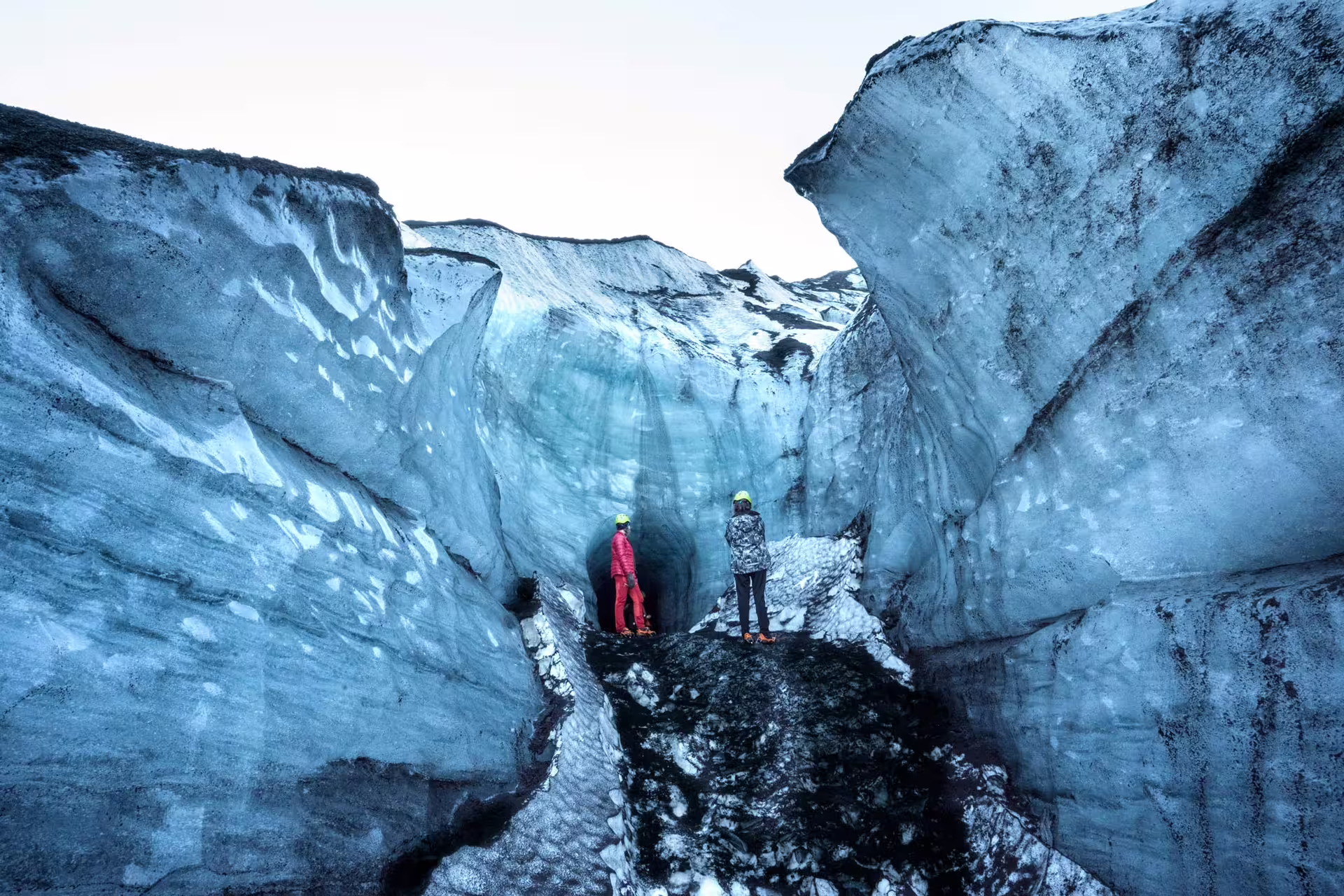 Two explorers in helmets standing inside Katla ice cave with towering blue glacial walls in Iceland