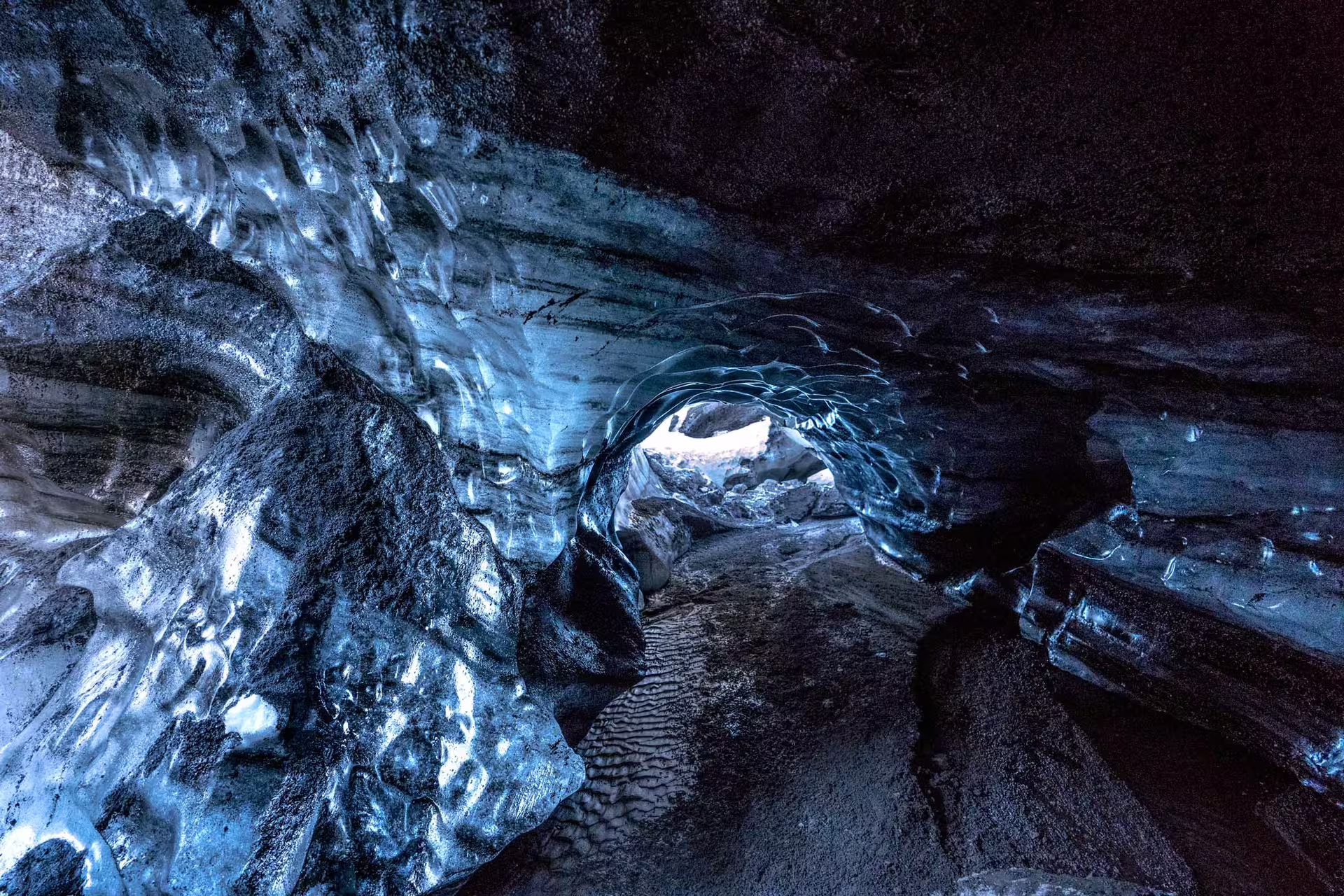 Blue glacial ice tunnel inside Katla ice cave with dark volcanic rock walls and light at the end