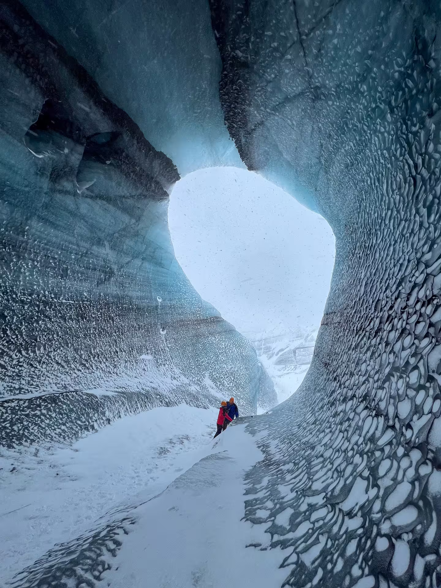 Two explorers inside Katla ice cave with dramatic textured glacier walls and bright cave opening in Iceland