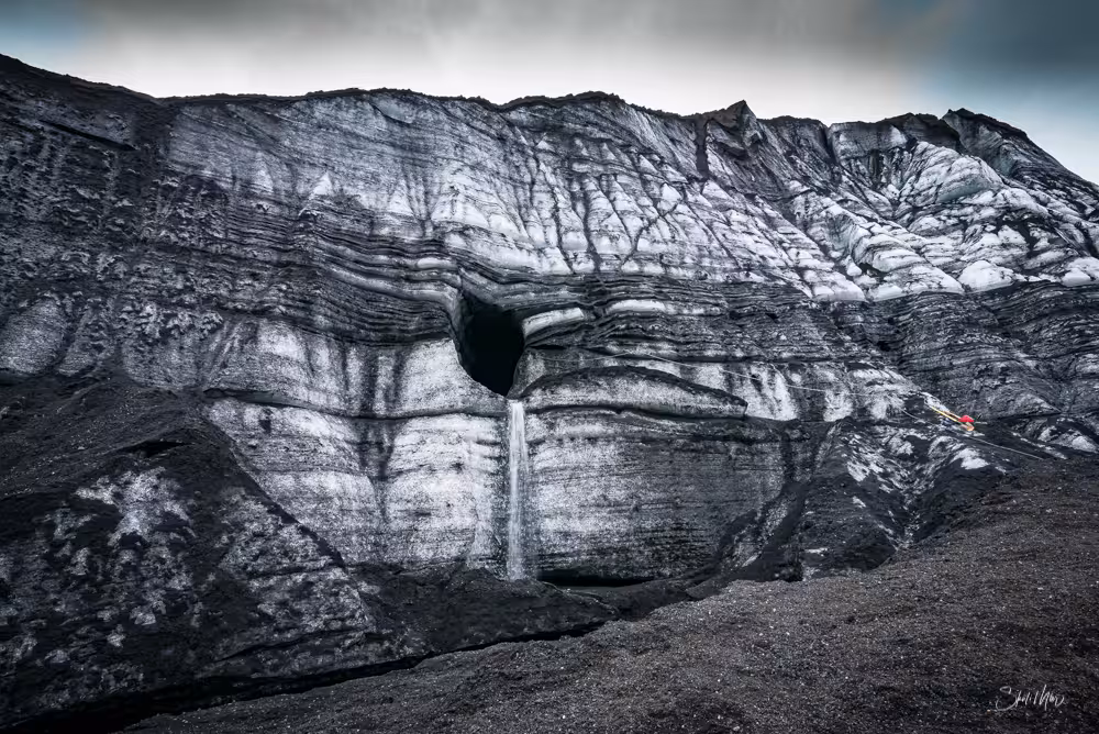 Dark layered glacier wall with cave opening and waterfall at Katla volcano Iceland
