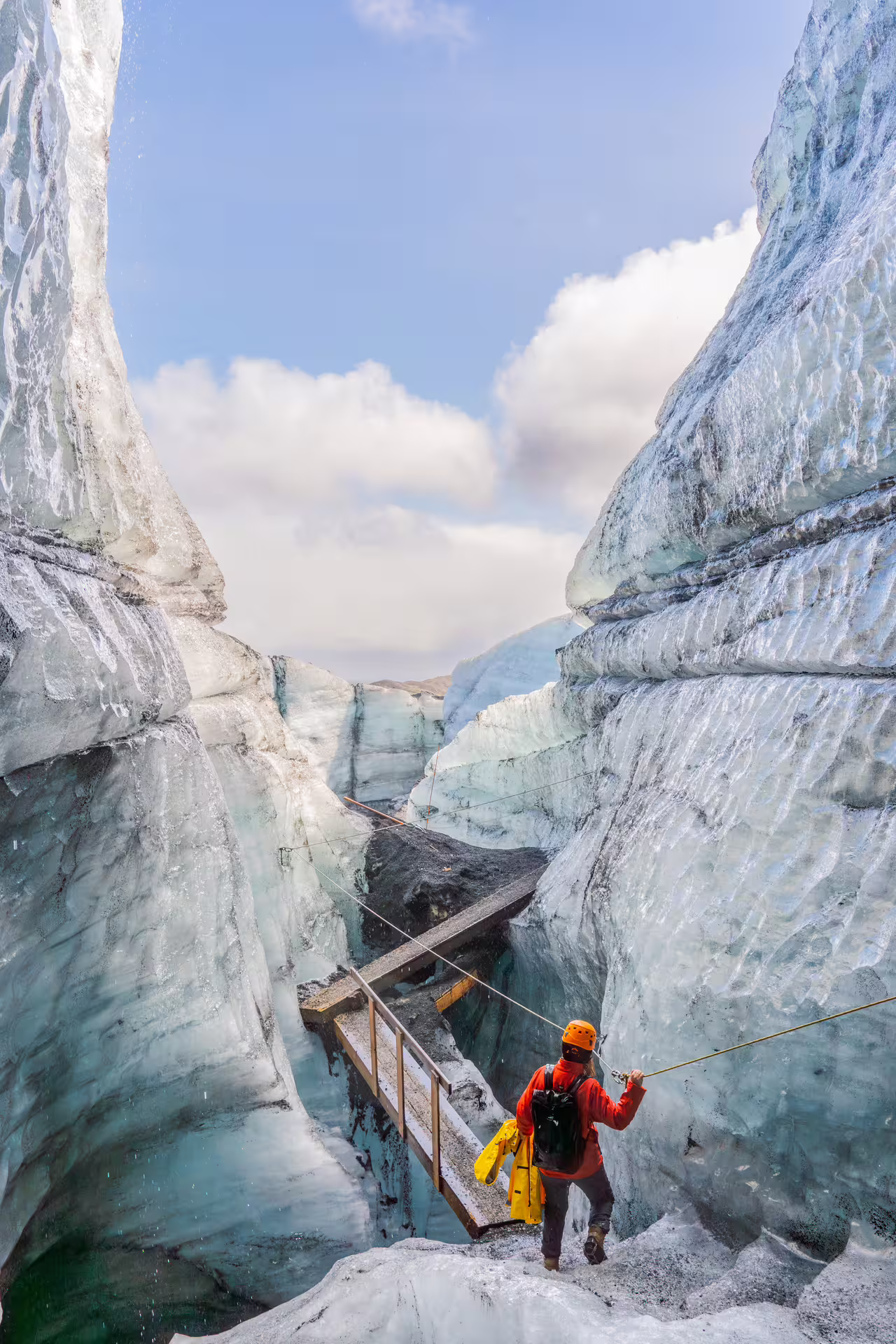 Hiker in orange gear crossing a wooden bridge through a narrow Katla ice cave canyon in Iceland
