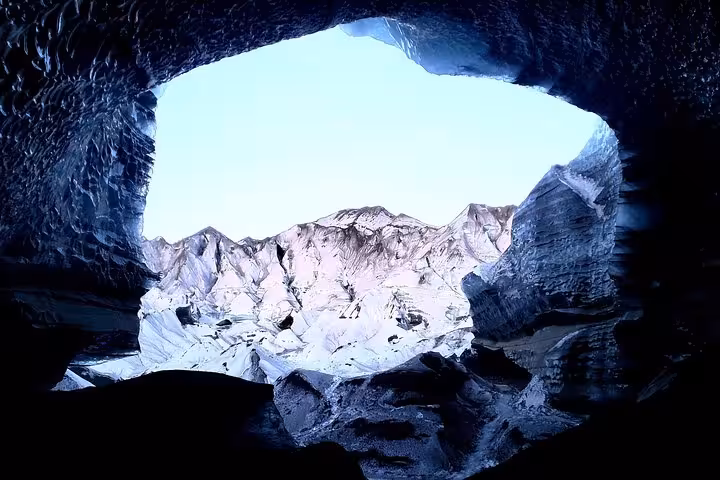 View from inside Katla ice cave showing snow-covered volcanic mountains through dark glacial cave opening