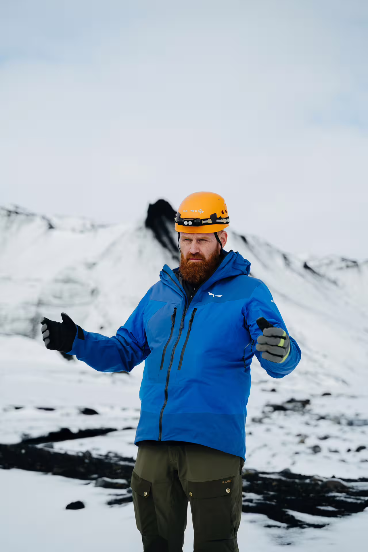 Tour guide in orange helmet and blue jacket gesturing in front of snowy Icelandic mountains