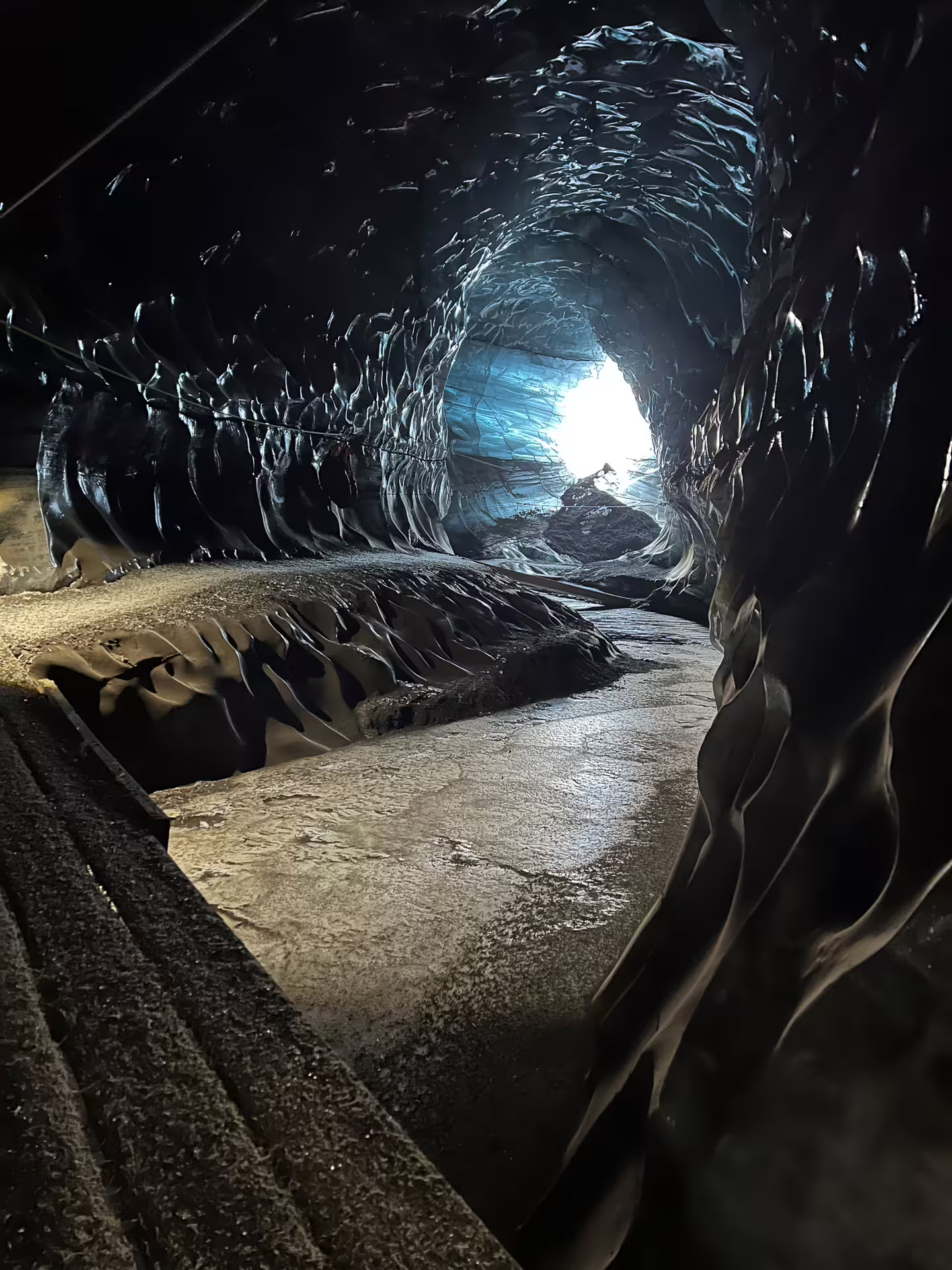 Dark glacial ice cave tunnel with black volcanic ash layers and glowing blue light at entrance, Katla Iceland
