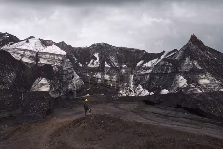 Hiker with yellow helmet standing before dramatic black volcanic mountains with white ice layers at Katla Iceland