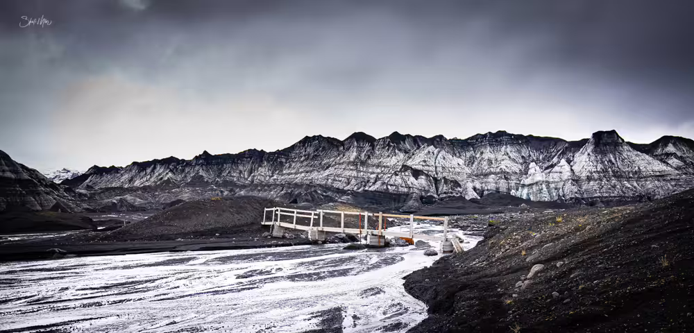 Black volcanic landscape with glacial river and small bridge near Katla, Iceland under dramatic stormy skies