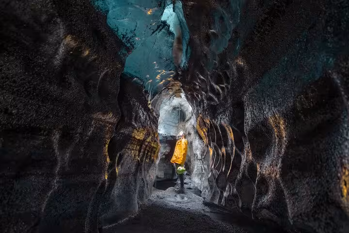 Explorer in yellow jacket standing inside dark Katla ice cave with volcanic black walls and turquoise glacier ceiling