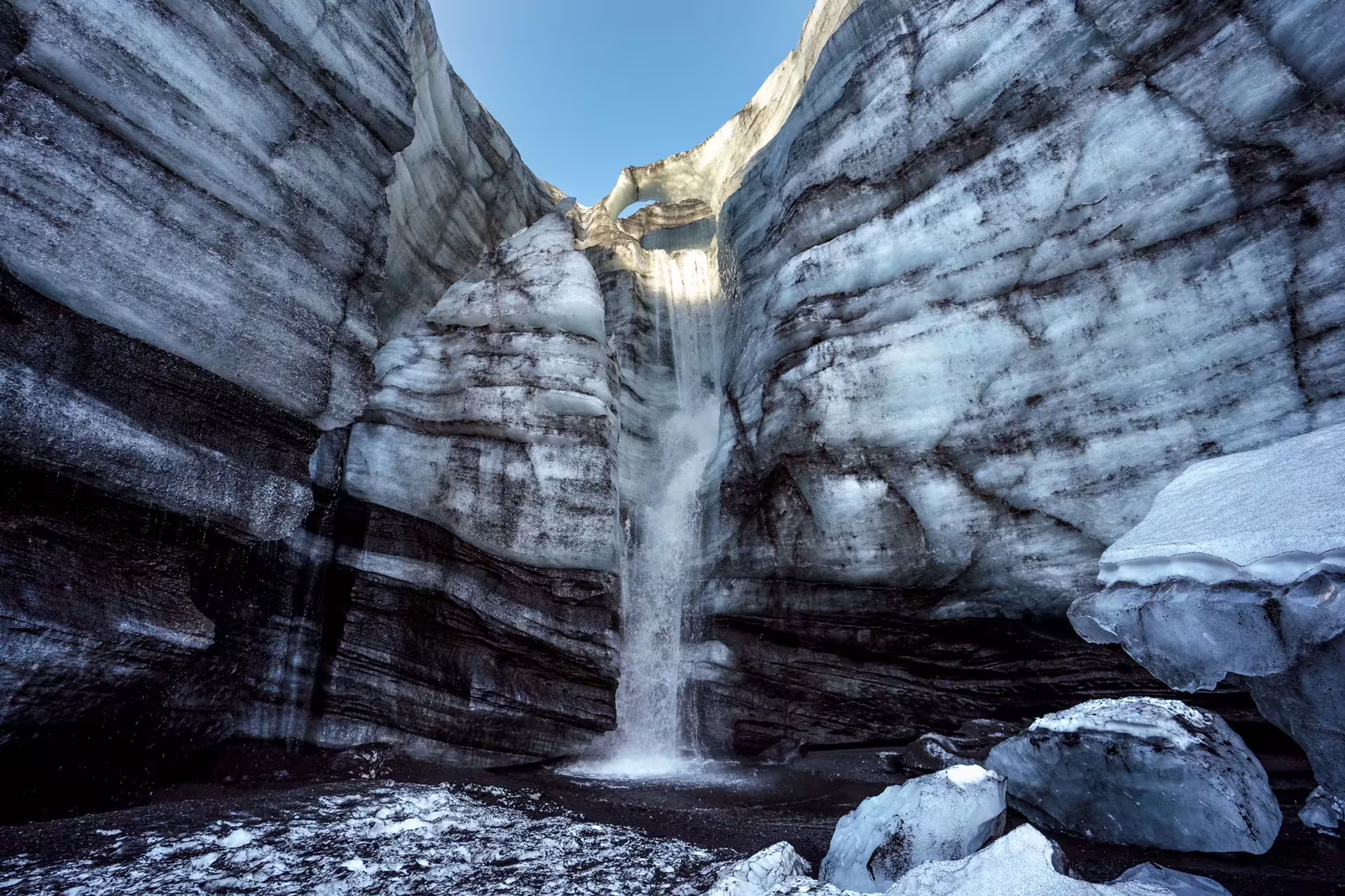 Meltwater waterfall cascading through dark layered ice canyon walls inside Katla ice cave Iceland