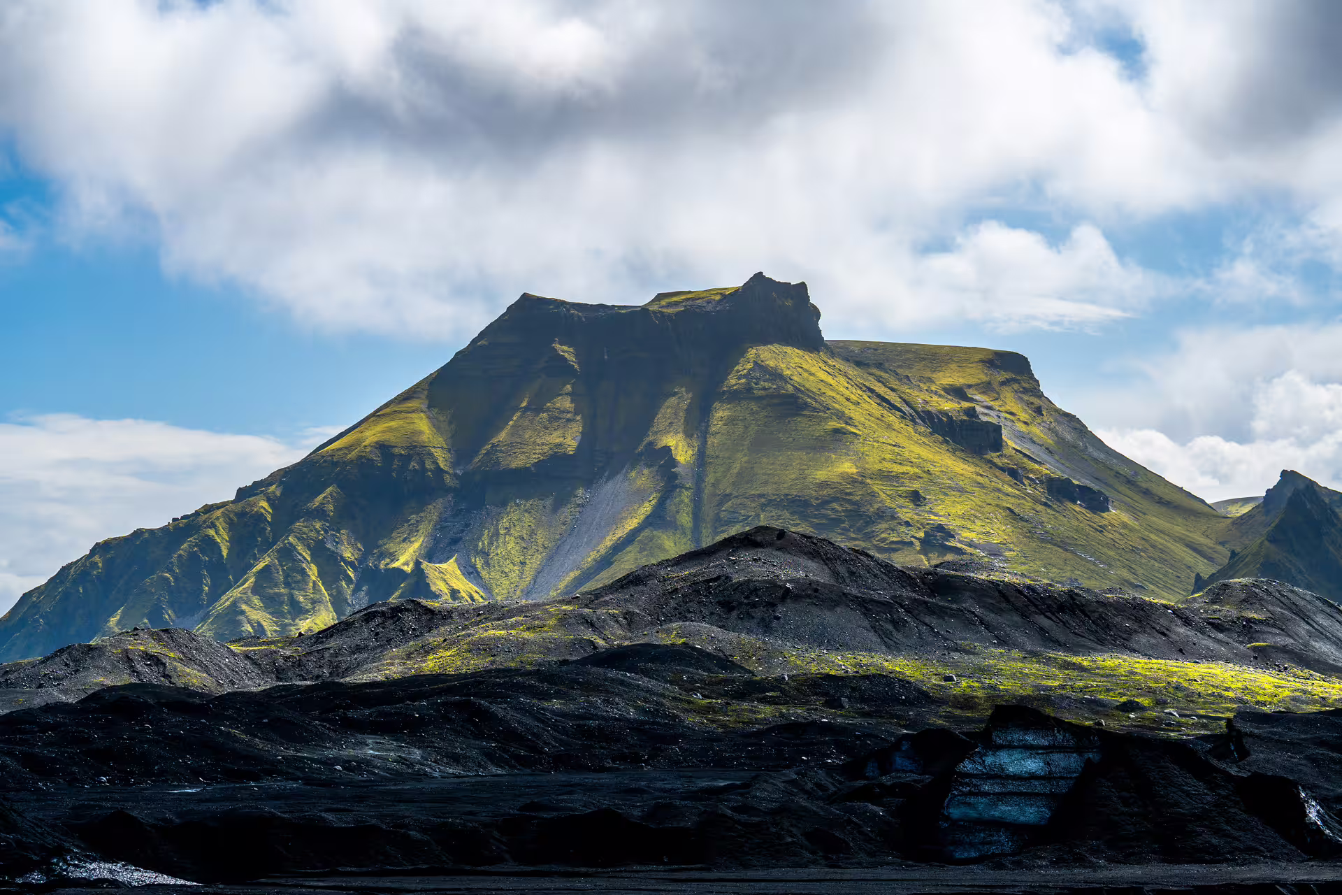 Black volcanic landscape with moss-covered mountain peak under blue sky near Katla, South Iceland