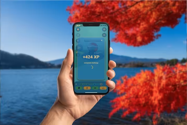 Hand holding smartphone showing +424 XP completed challenge on GPS walking tour app at Lake Kawaguchiko with autumn foliage
