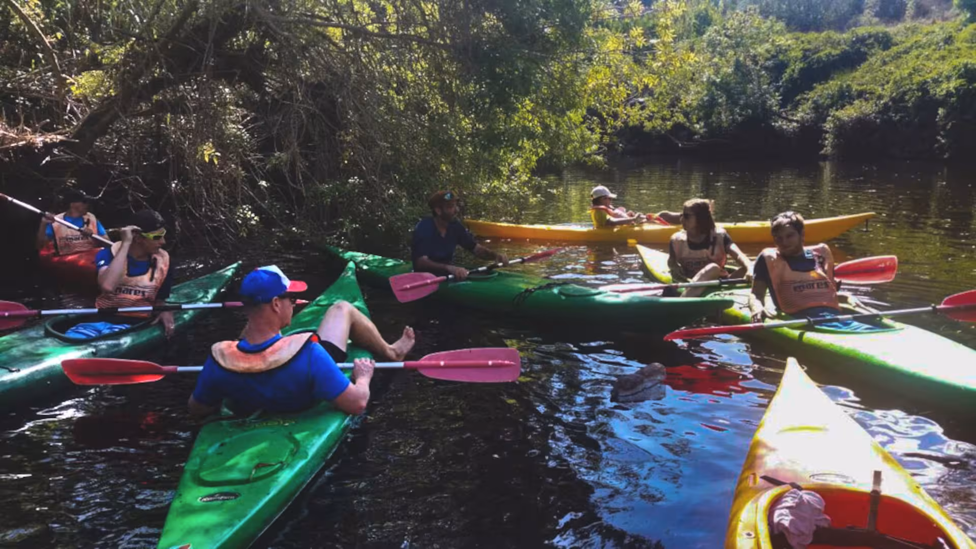 Kayakers gathered on the tranquil Temo River, Bosa, surrounded by vibrant nature and shaded by trees.