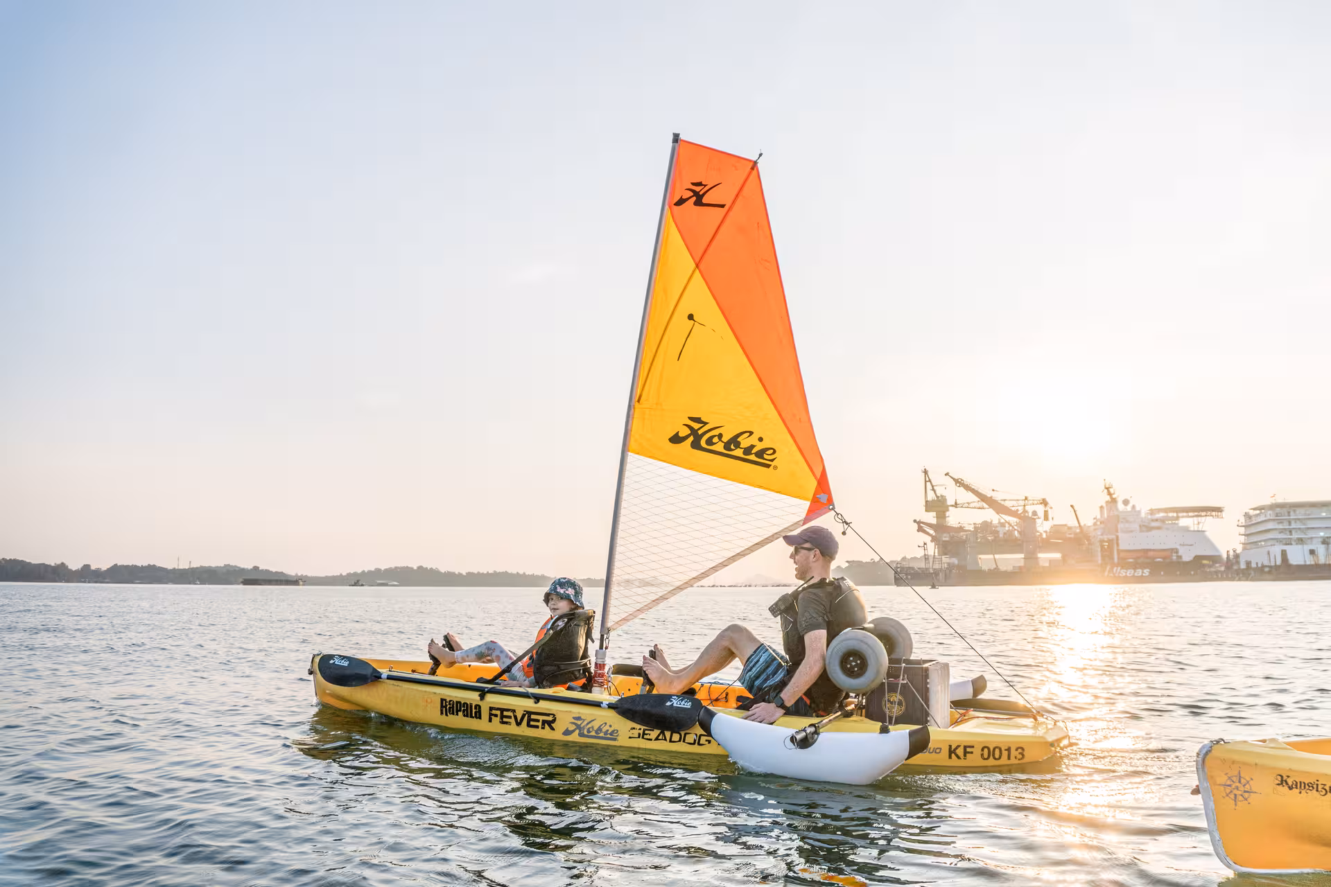 Two adventurers enjoy a sunset kayak sailing adventure near a bustling harbor with vibrant sails.