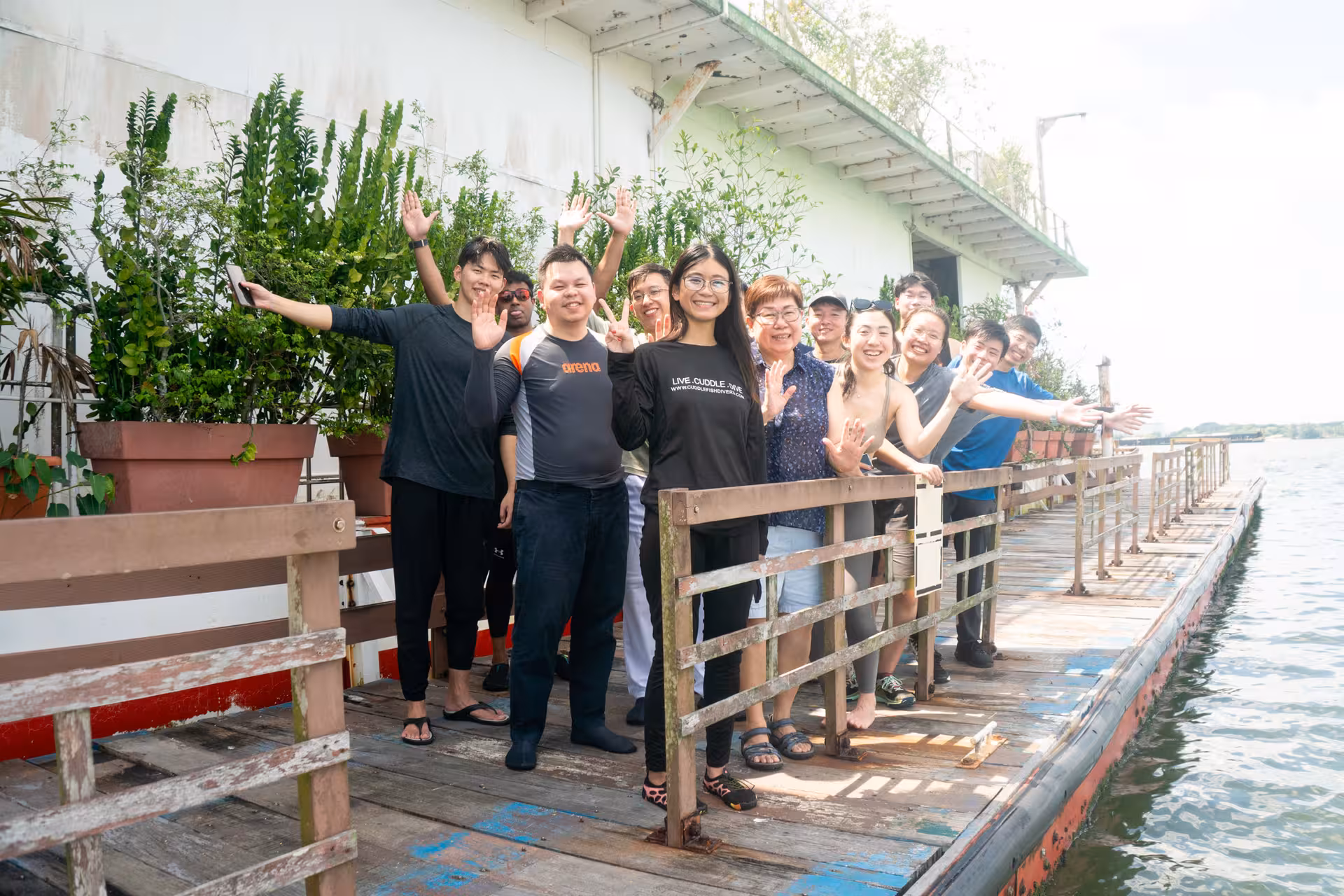 Group of happy tourists waving on a dock, ready for their kayak sailing adventure by the water's edge.