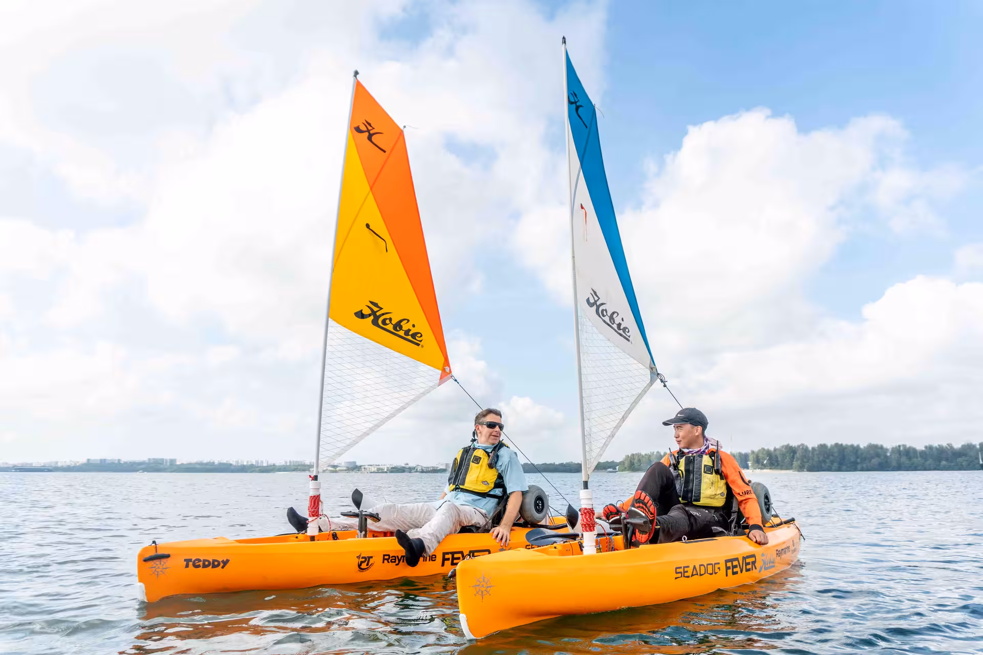 Two cheerful adventurers enjoy a sunny day on the water in vibrant sail-equipped kayaks.