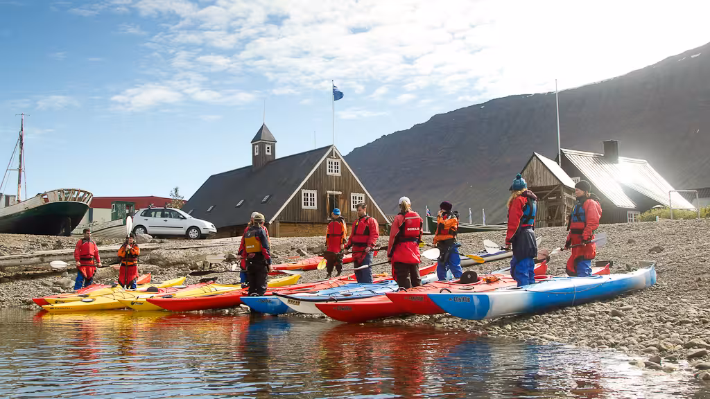 Kayakers in drysuits gather by colorful sea kayaks at a fjord shoreline base before a calm water paddling tour