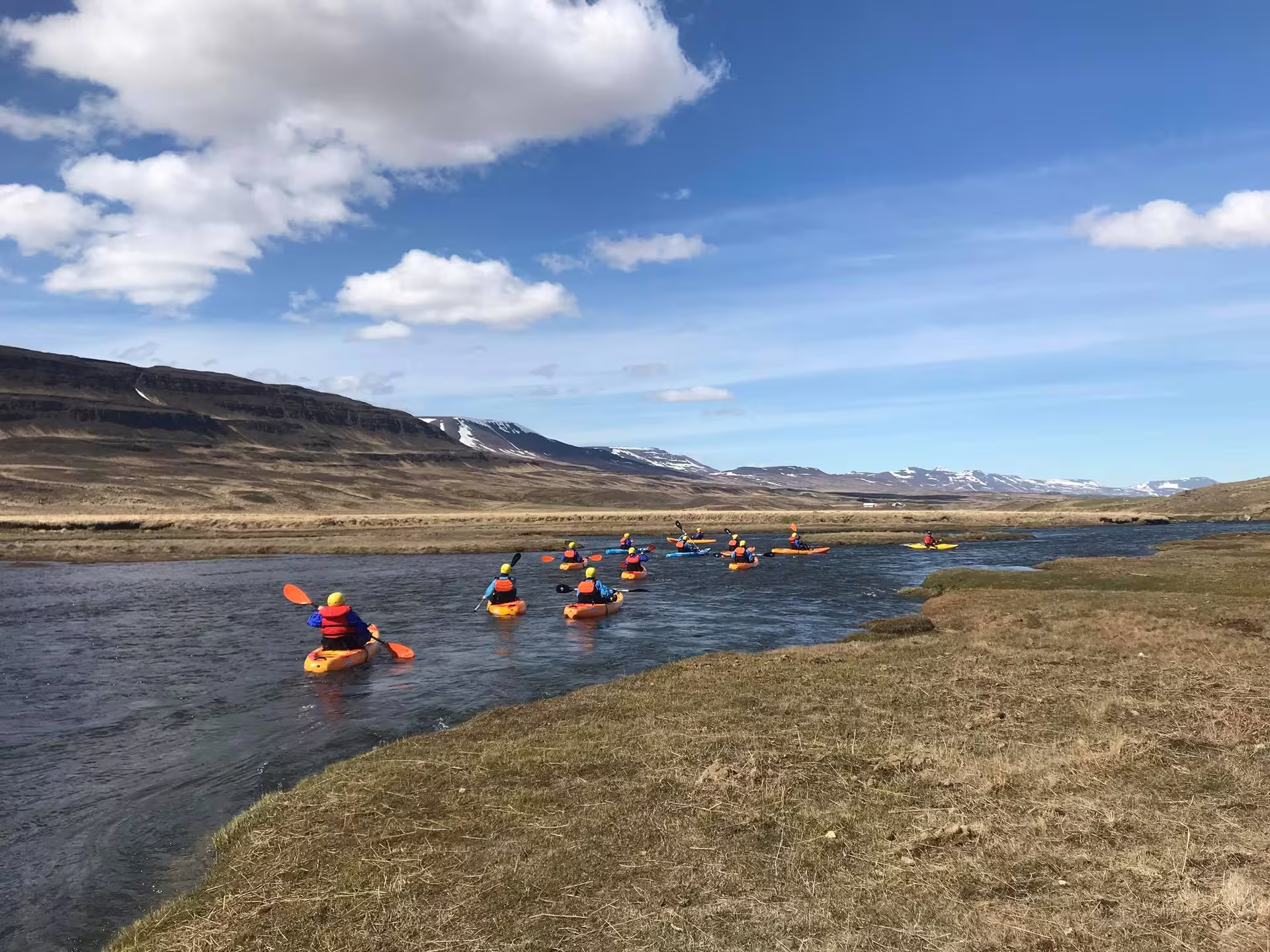 Kayakers paddle down the scenic Svartá River under a blue sky, surrounded by Iceland's stunning mountainous landscape.