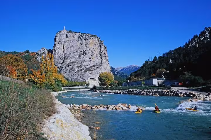 Kayakers paddling on turquoise river beside towering limestone rock with chapel at Castellane, Gorges du Verdon