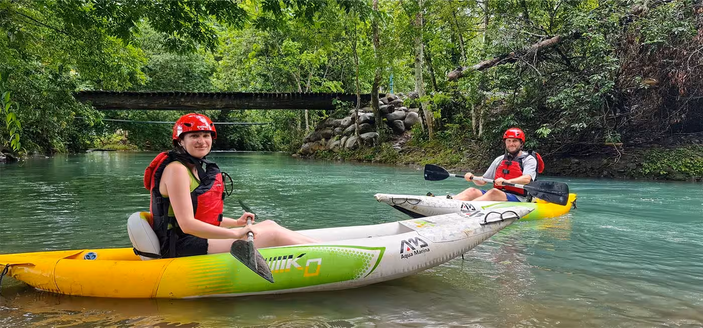 Two kayakers in bright helmets paddle through lush greenery on Río Frío, showcasing adventure and nature exploration.