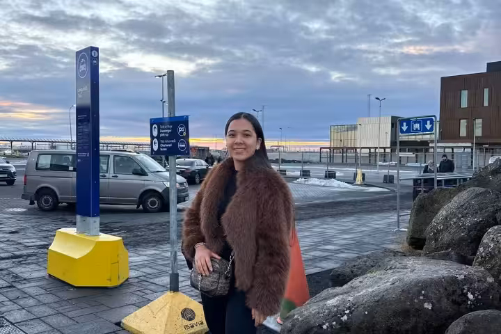 Woman in fur coat standing outside Keflavik Airport Iceland near parking signs at sunset