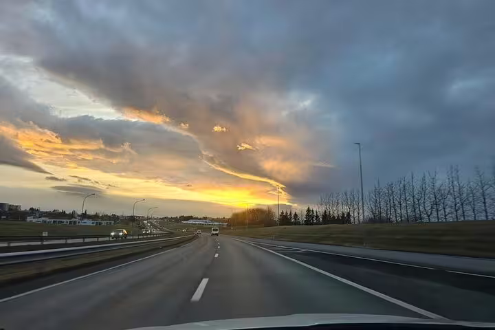 Highway drive at sunset toward Reykjavik with dramatic orange clouds and sparse trees along roadside