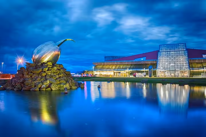 Keflavik Airport terminal at dusk with illuminated sculpture reflected in calm water