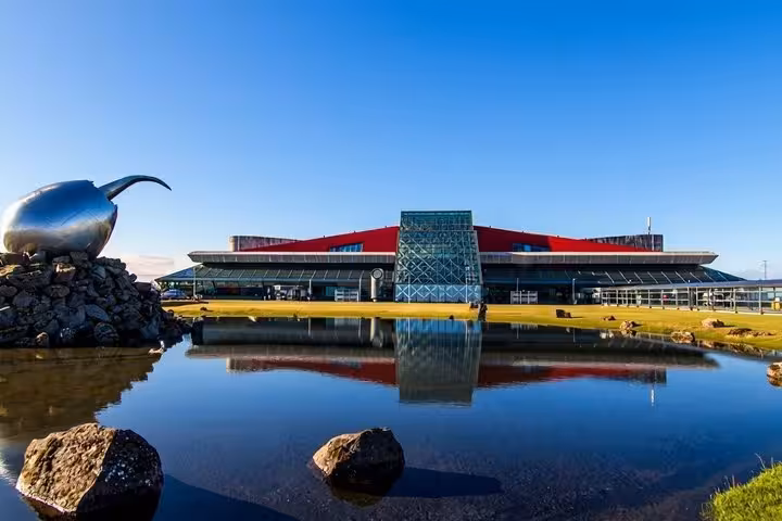 Keflavik International Airport terminal with red roof, glass facade, and reflective pond under clear blue sky