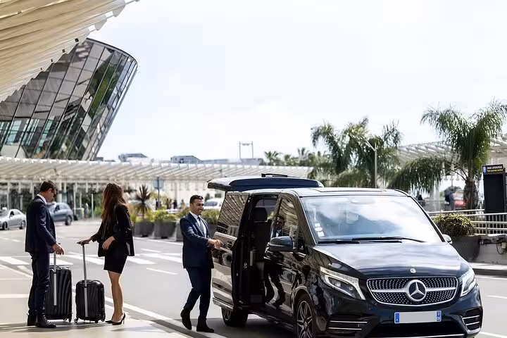Chauffeur opening door of black Mercedes V-Class van for two passengers with luggage at airport