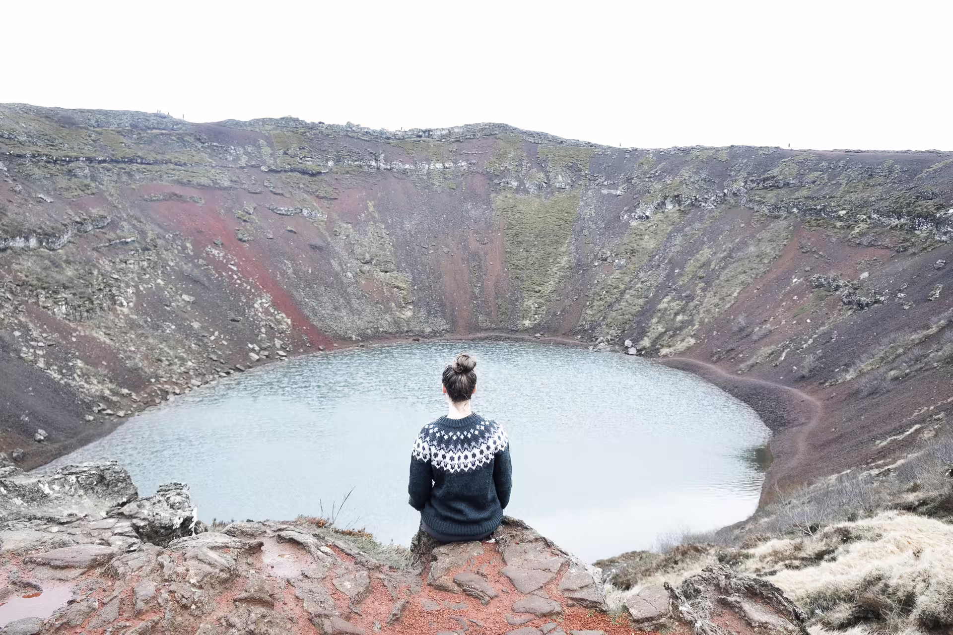 Person in Icelandic sweater sitting on volcanic rim overlooking Kerid crater lake on Golden Circle route
