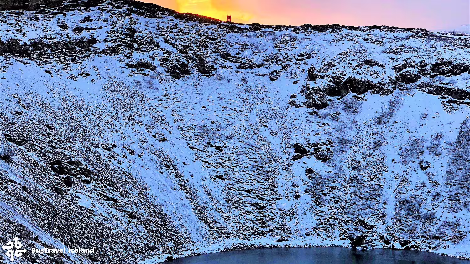Snow-covered Kerid volcano crater with frozen lake at sunset, two silhouetted figures on rim, Iceland