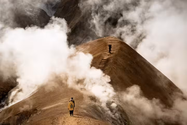 Hiker in yellow jacket walking geothermal ridge at Kerlingarfjoll Highlands with steam vents and glacier backdrop