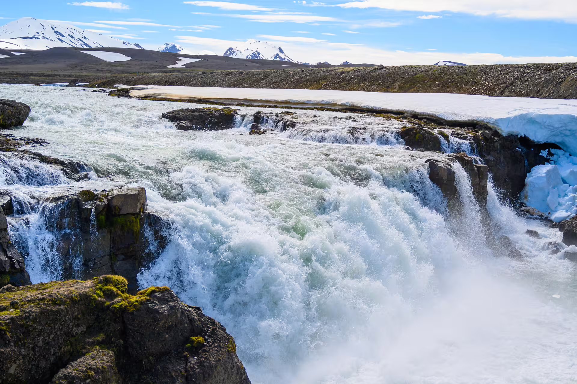 Powerful glacial waterfall cascading over rocky ledge with snow-covered highlands and mountains in background, Iceland